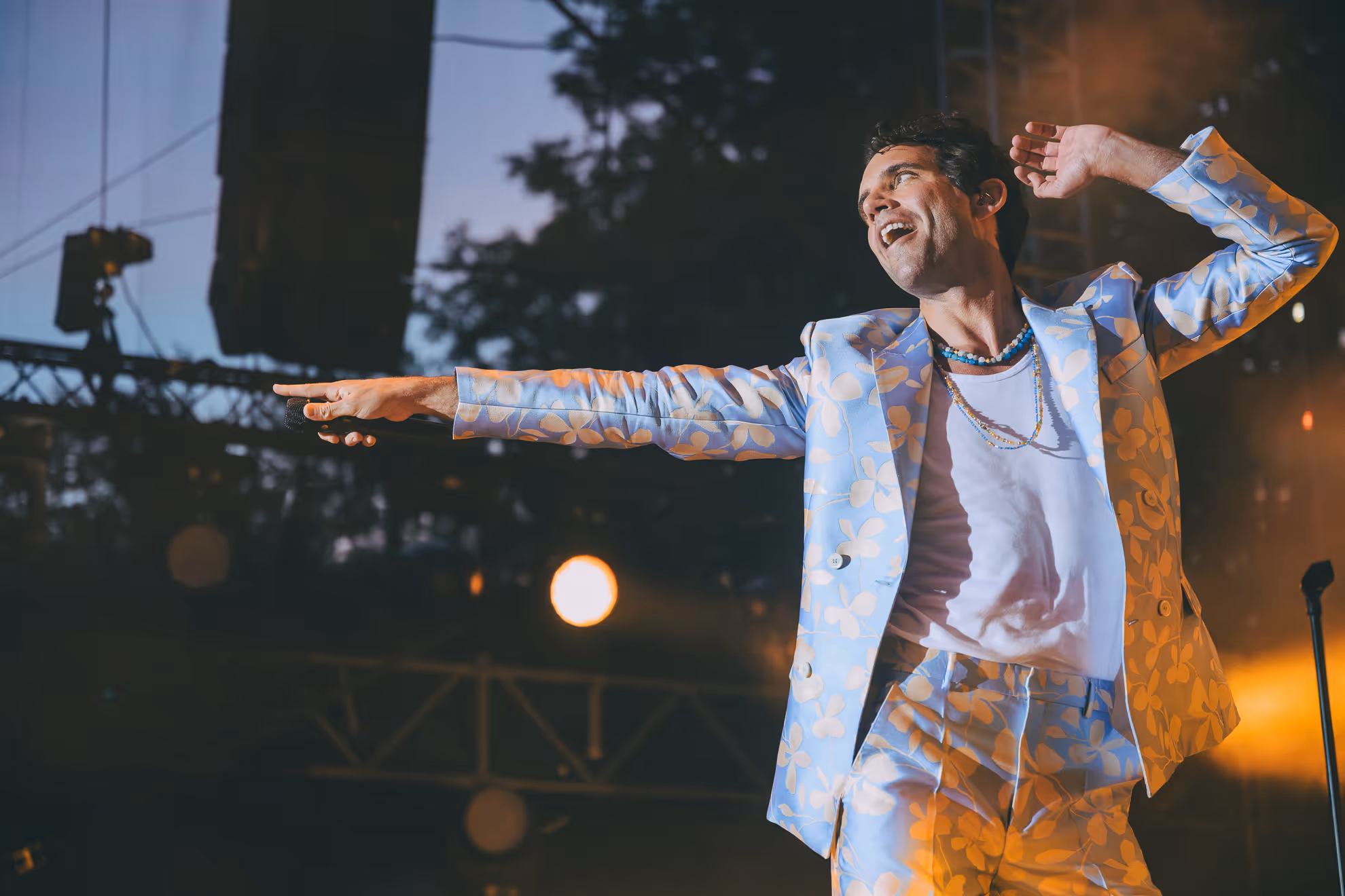 Homme souriant avec chemise blanche à motifs, sous des lumières de scène lors d'un concert en soirée.