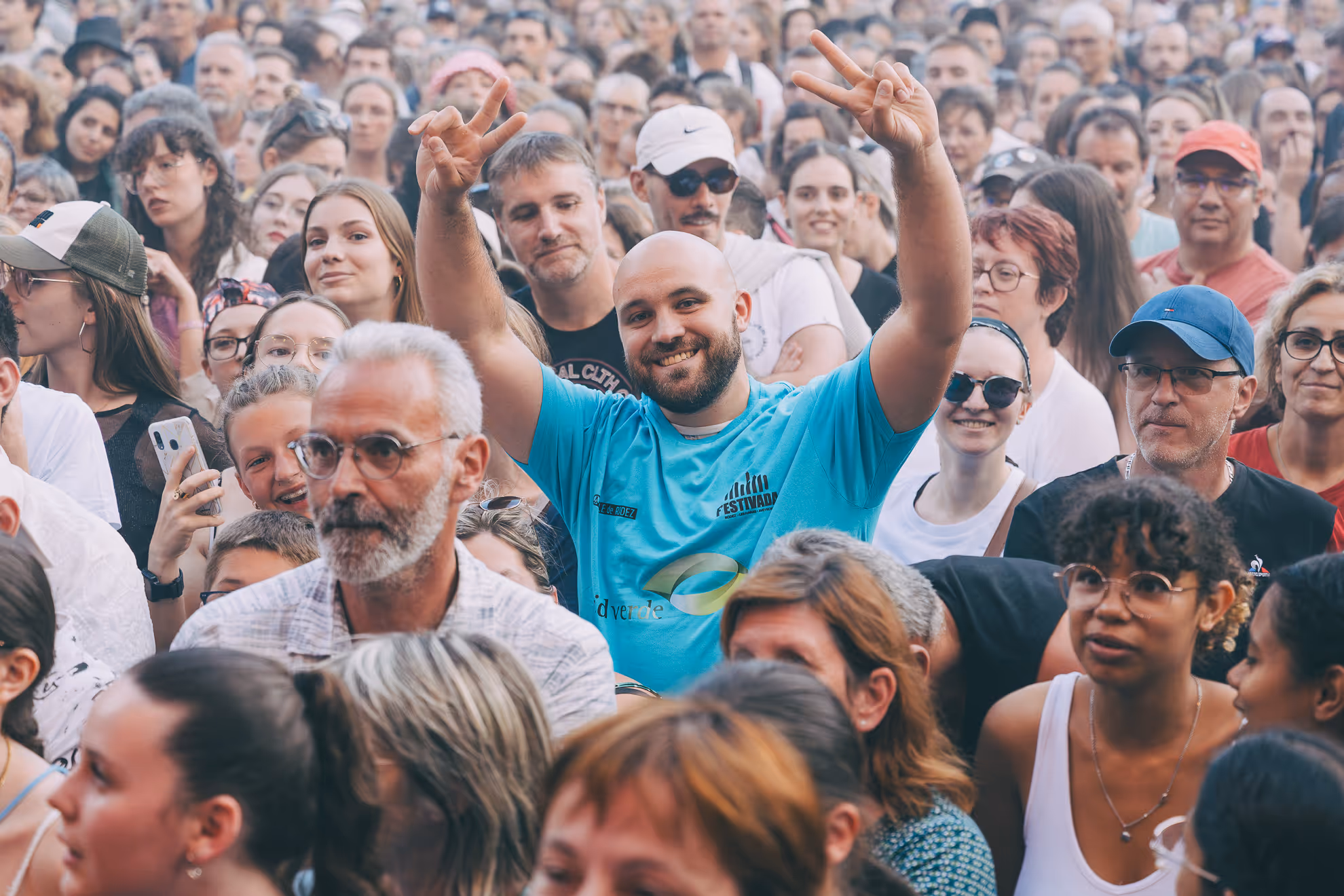 Une foule diverse et nombreuse lors d'un événement, avec un homme souriant au centre levant les deux mains en signe de paix.