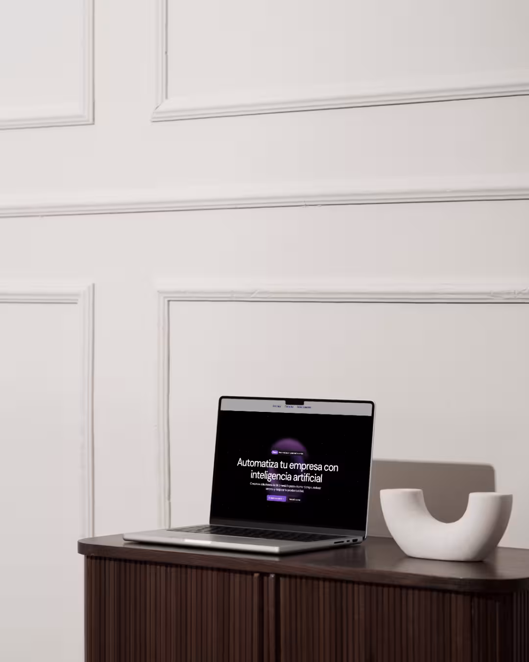 Portátil abierto sobre mueble oscuro con texto en pantalla que dice 'Automatiza tu empresa con inteligencia artificial' y una figura decorativa blanca al lado.