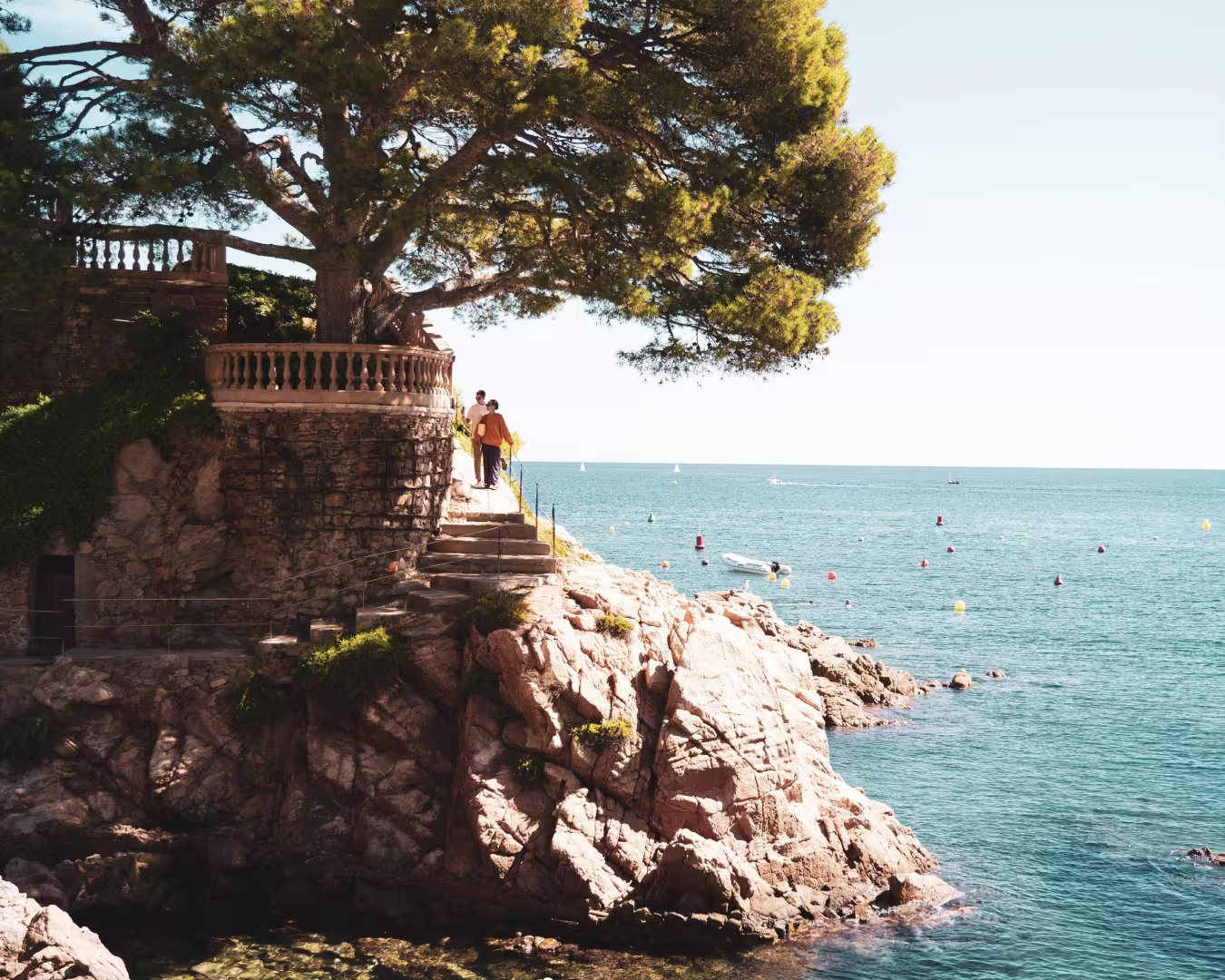 Two people walking up stone stairs along a rocky cliff by the sea with a large tree overhead and boats in the water.
