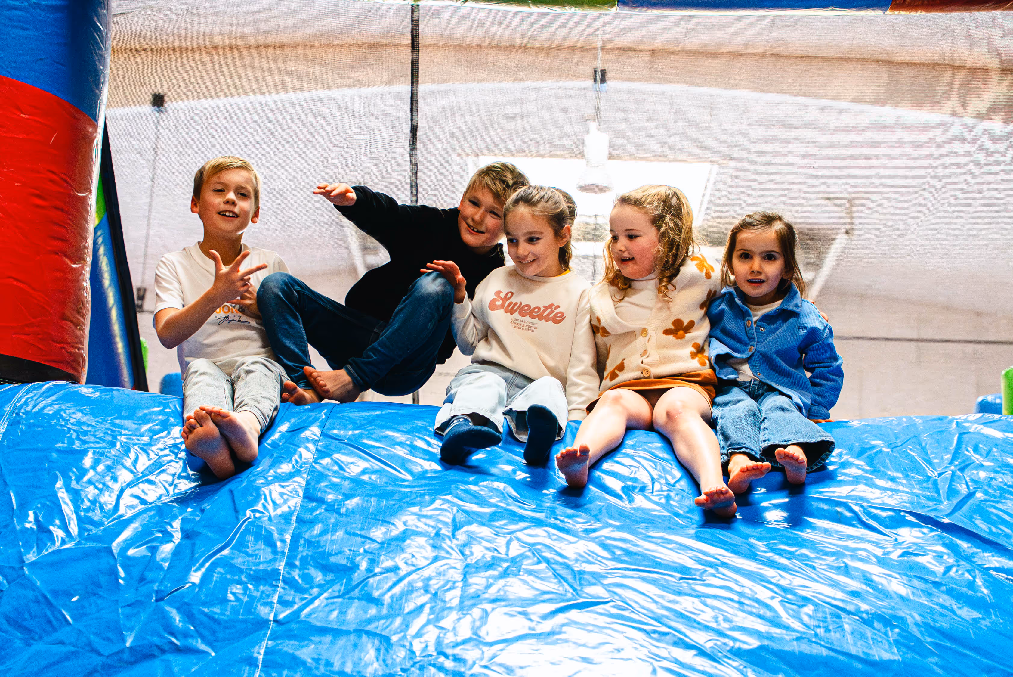 Drie meisjes en één jongen zitten boven op een blauw springkasteel en bereiden zich voor om samen naar beneden te glijden, in een lichte binnenruimte.