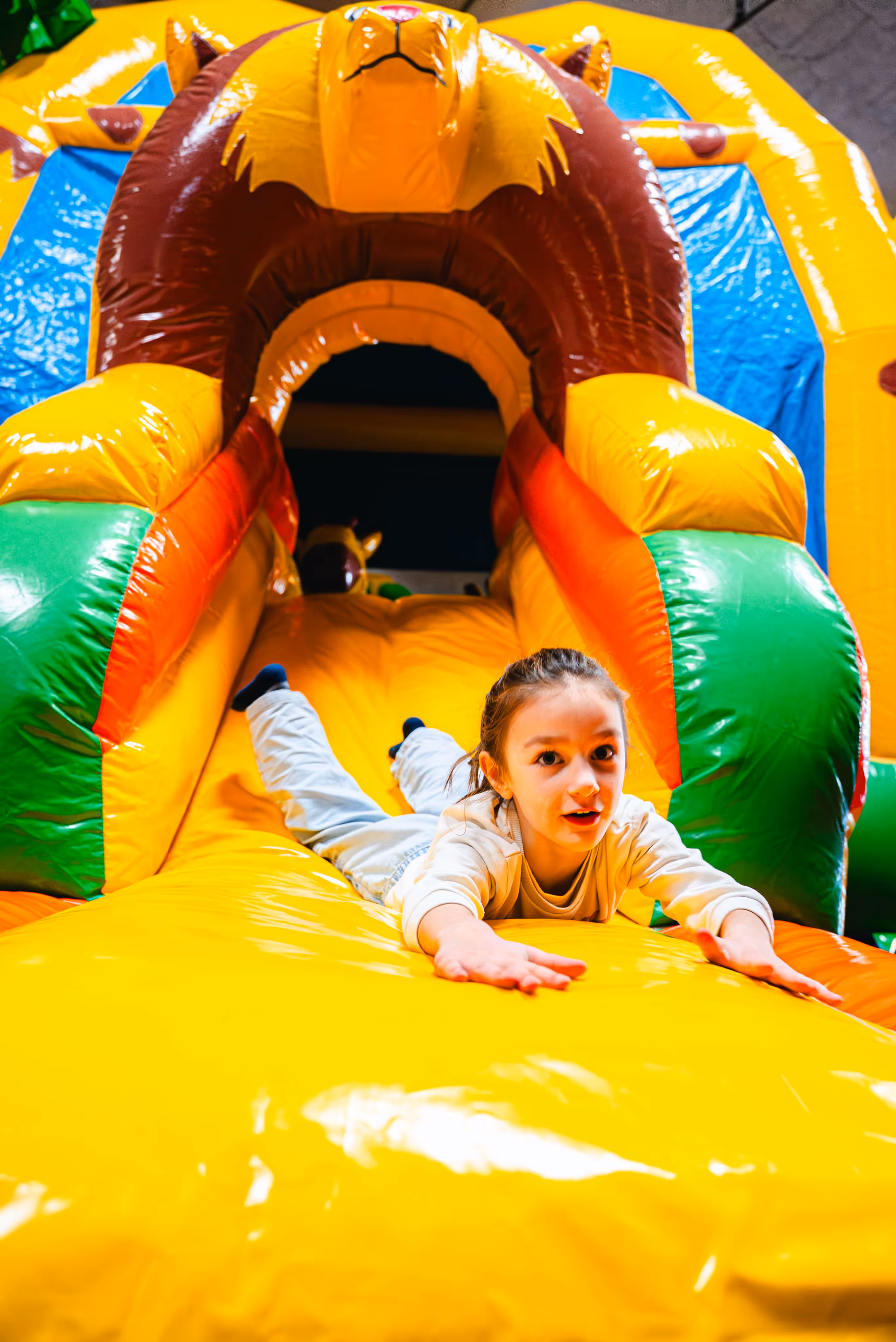 Jong meisje glijdt enthousiast naar beneden van een geel springkasteel met kleurrijke boogvormige ingang, in een speelse binnenruimte.