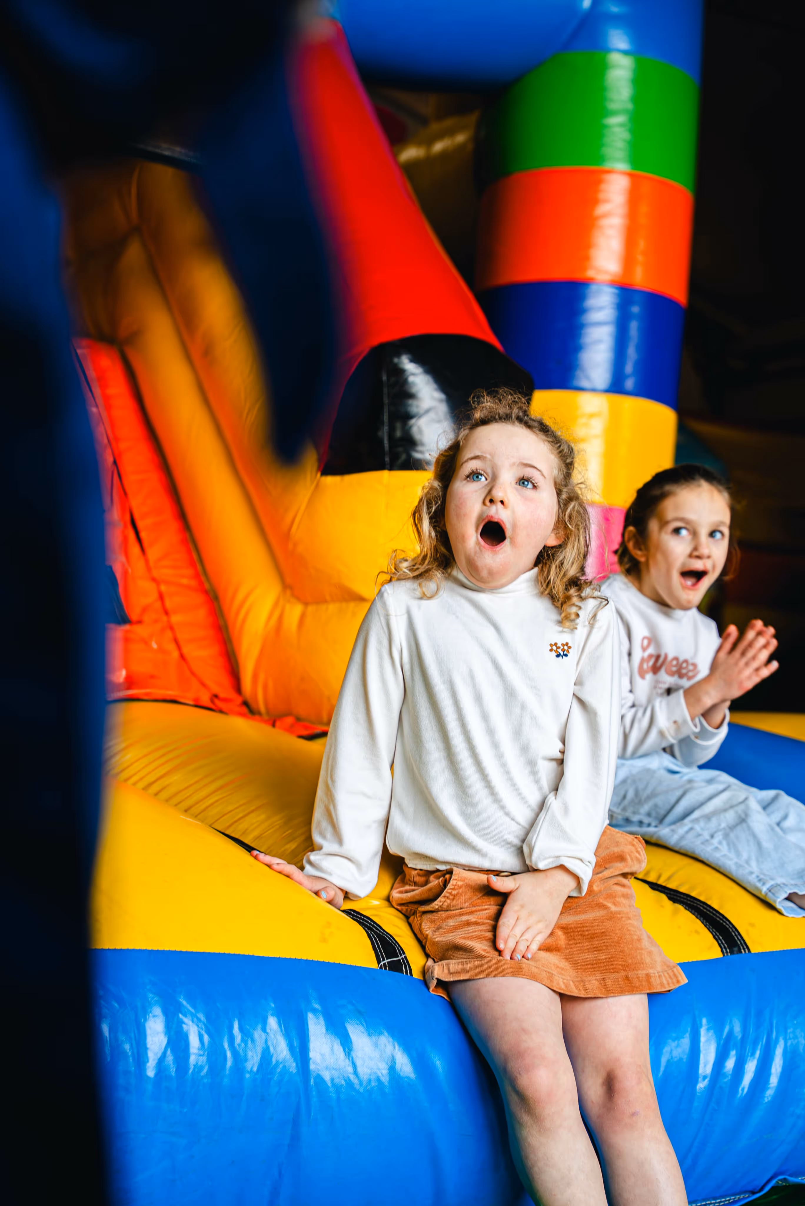 Twee kinderen zitten op een kleurrijk springkasteel, met een verbaasde blik en open mond.