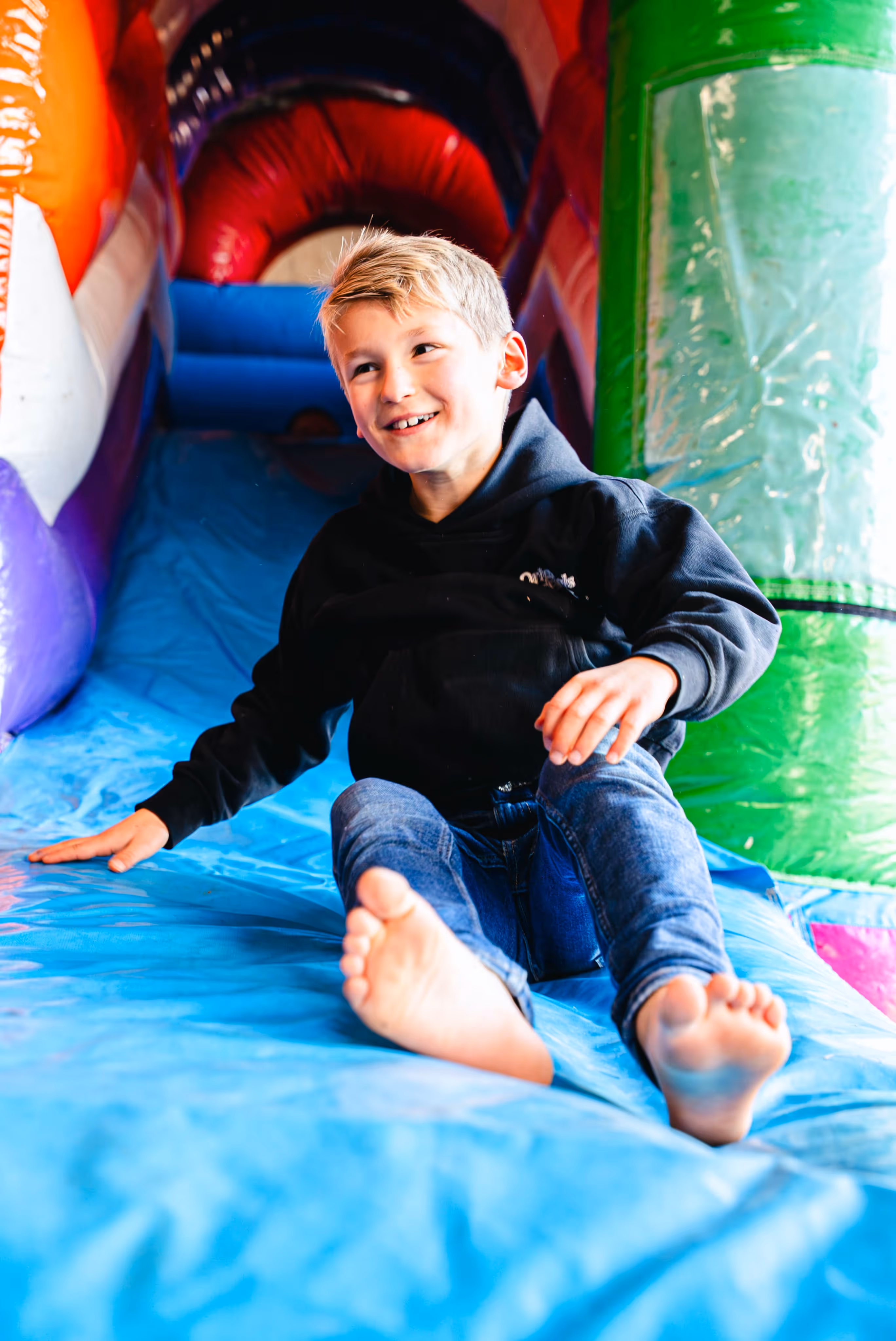Jonge jongen zit lachend onderaan een blauwe glijbaan van een kleurrijk springkasteel, in een speelse binnenruimte.