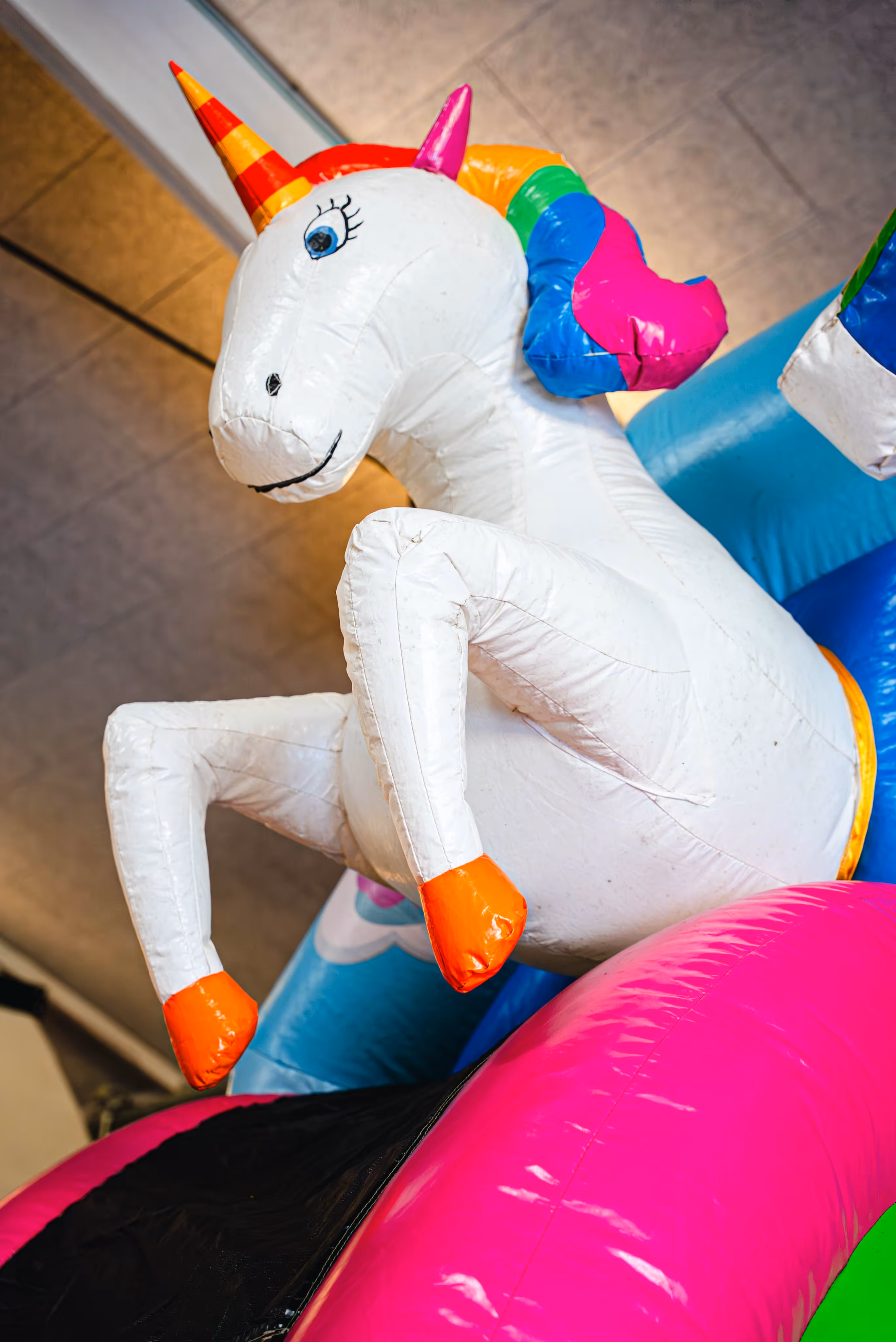 Detail van een eenhoorn-thema springkasteel met een grote witte opblaasbare eenhoorn met gekleurde hoorn en manen, staand op een felgekleurde glijbaan in een binnenruimte.