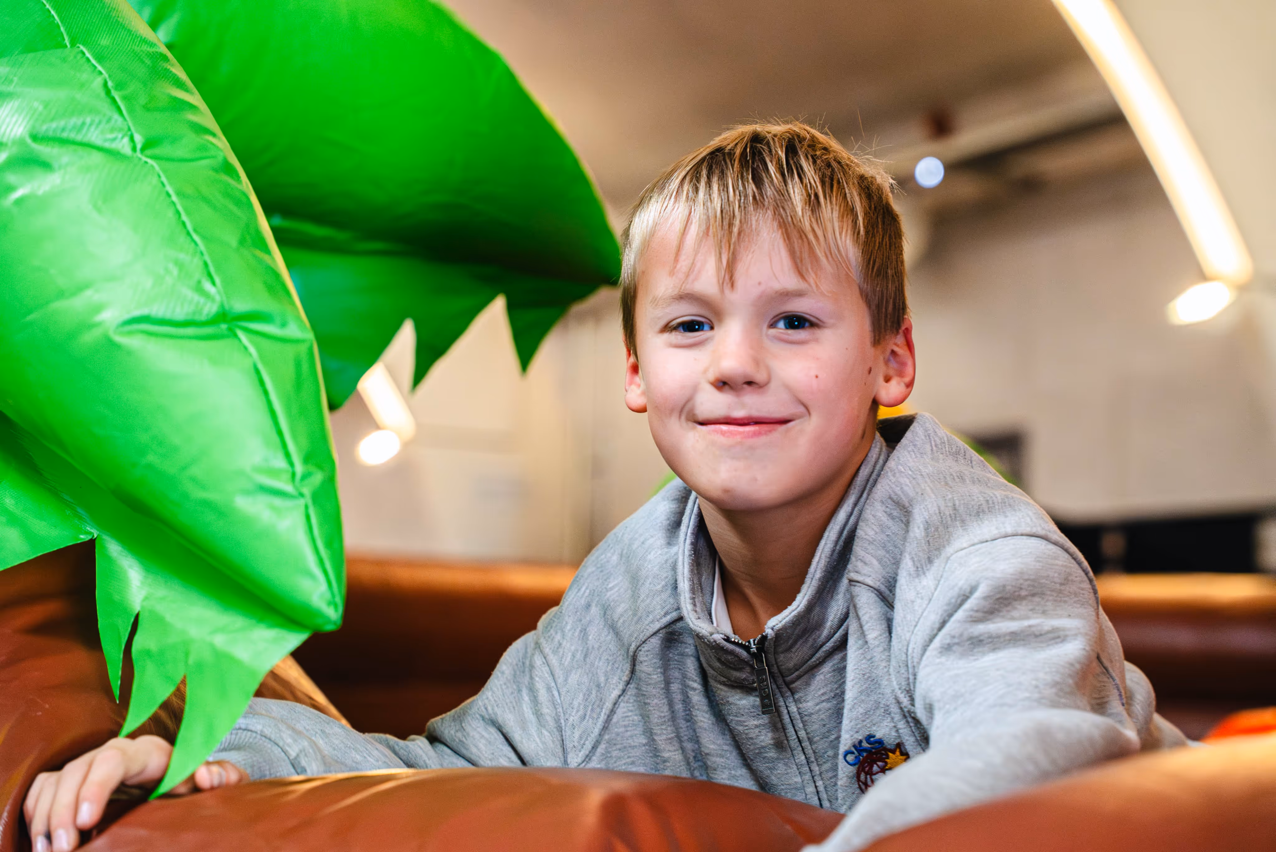 Jonge jongen kijkt lachend in de camera terwijl hij speelt bij een groen jungle-element van een springkasteel, in een lichte binnenruimte.