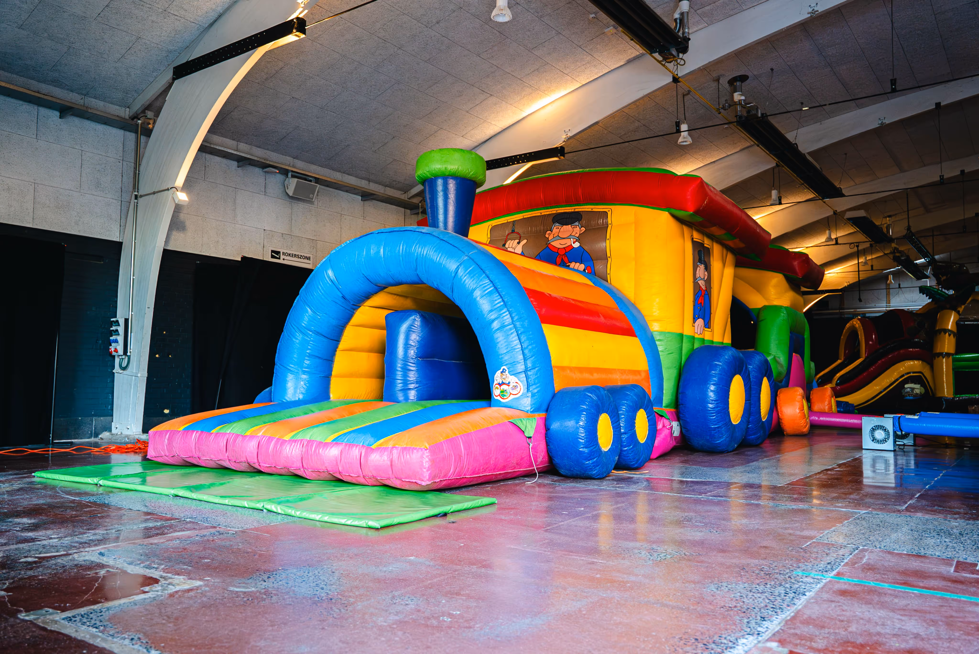 Groot, kleurrijk trein-thema springkasteel met meerdere wagons en een boogvormige ingang, opgesteld in een ruime binnenhal.