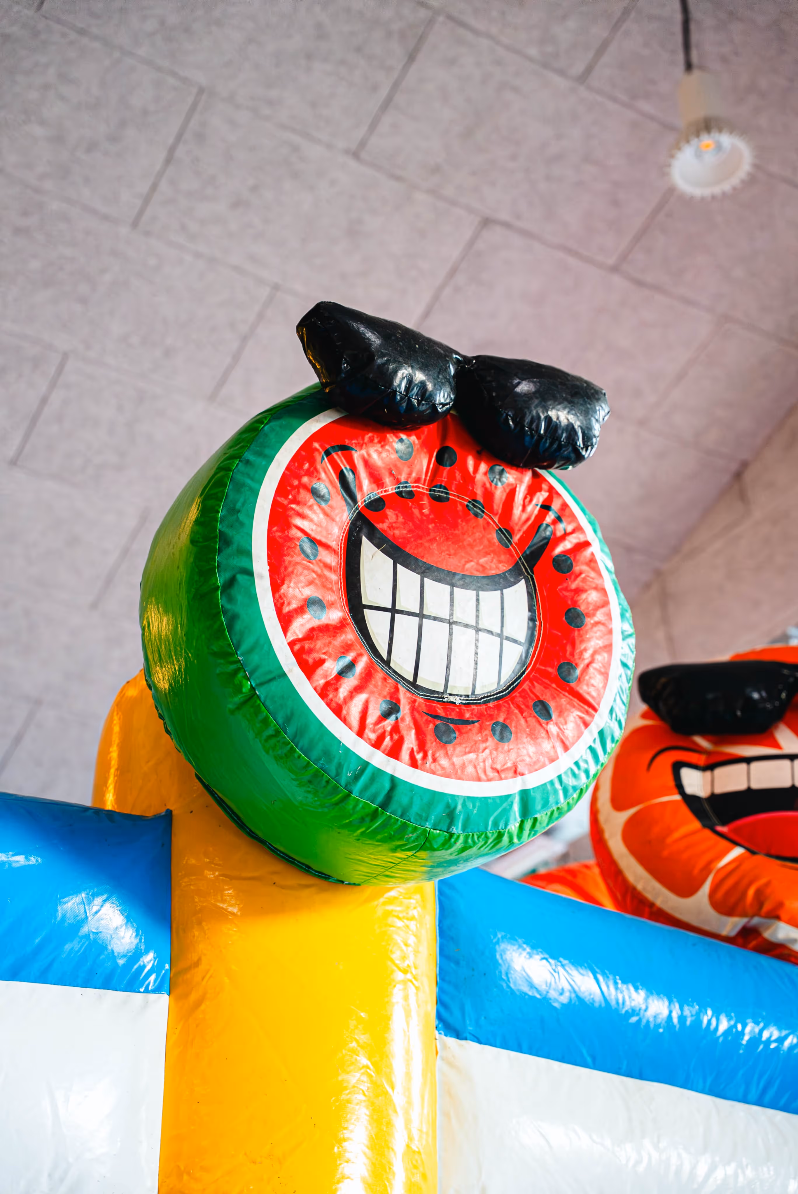 Detail van een kleurrijk springkasteel met een ronde, watermeloenachtige vorm, een grote lachende mond met witte tanden en zwarte cartoonogen.