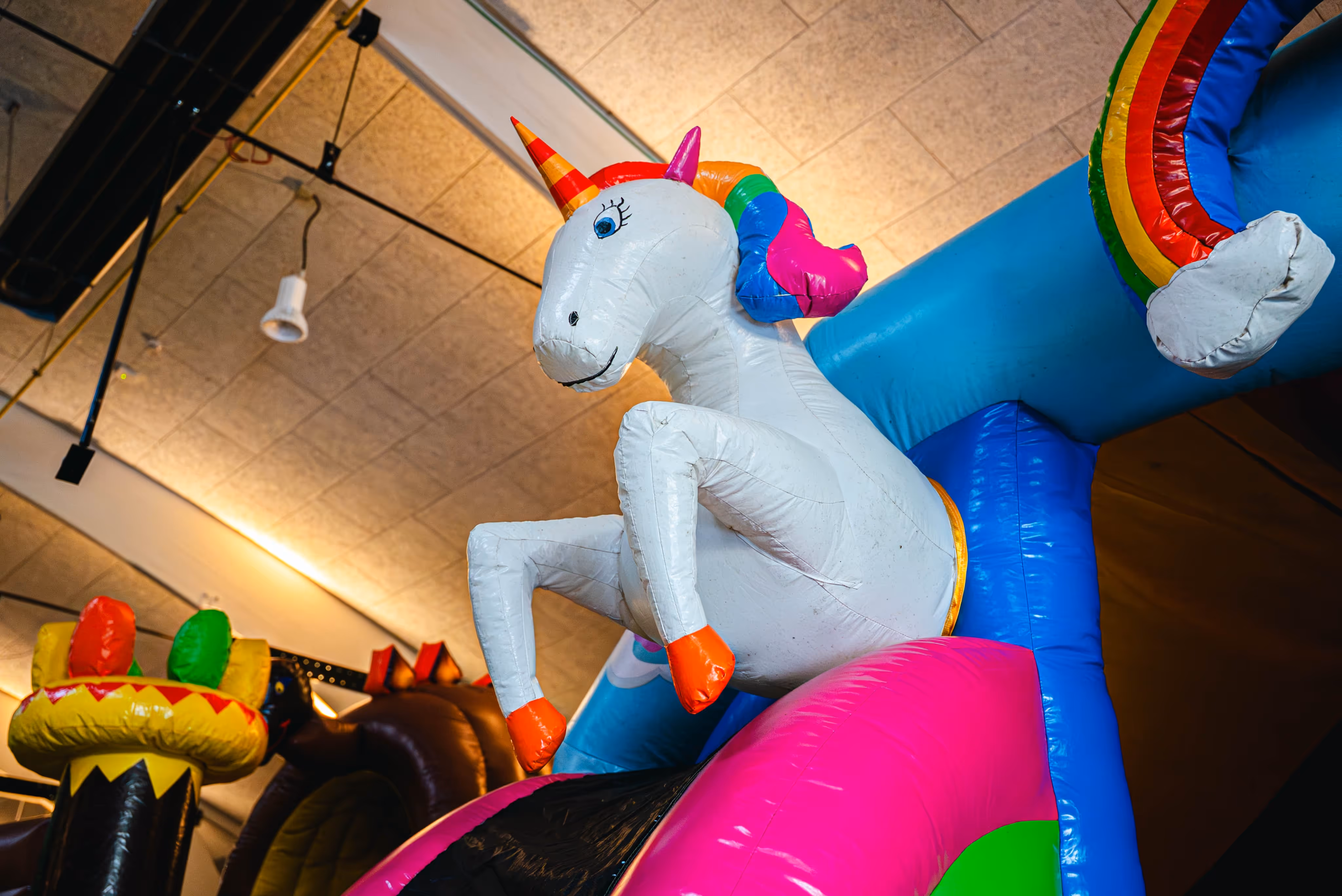 Detail van een eenhoorn-thema springkasteel met een grote witte opblaasbare eenhoorn met gekleurde hoorn en manen, staand op een felgekleurde glijbaan in een binnenruimte.