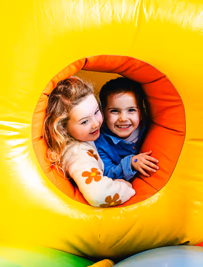 Twee jonge meisjes lachen samen door een ronde opening van een geel en oranje springkasteel.