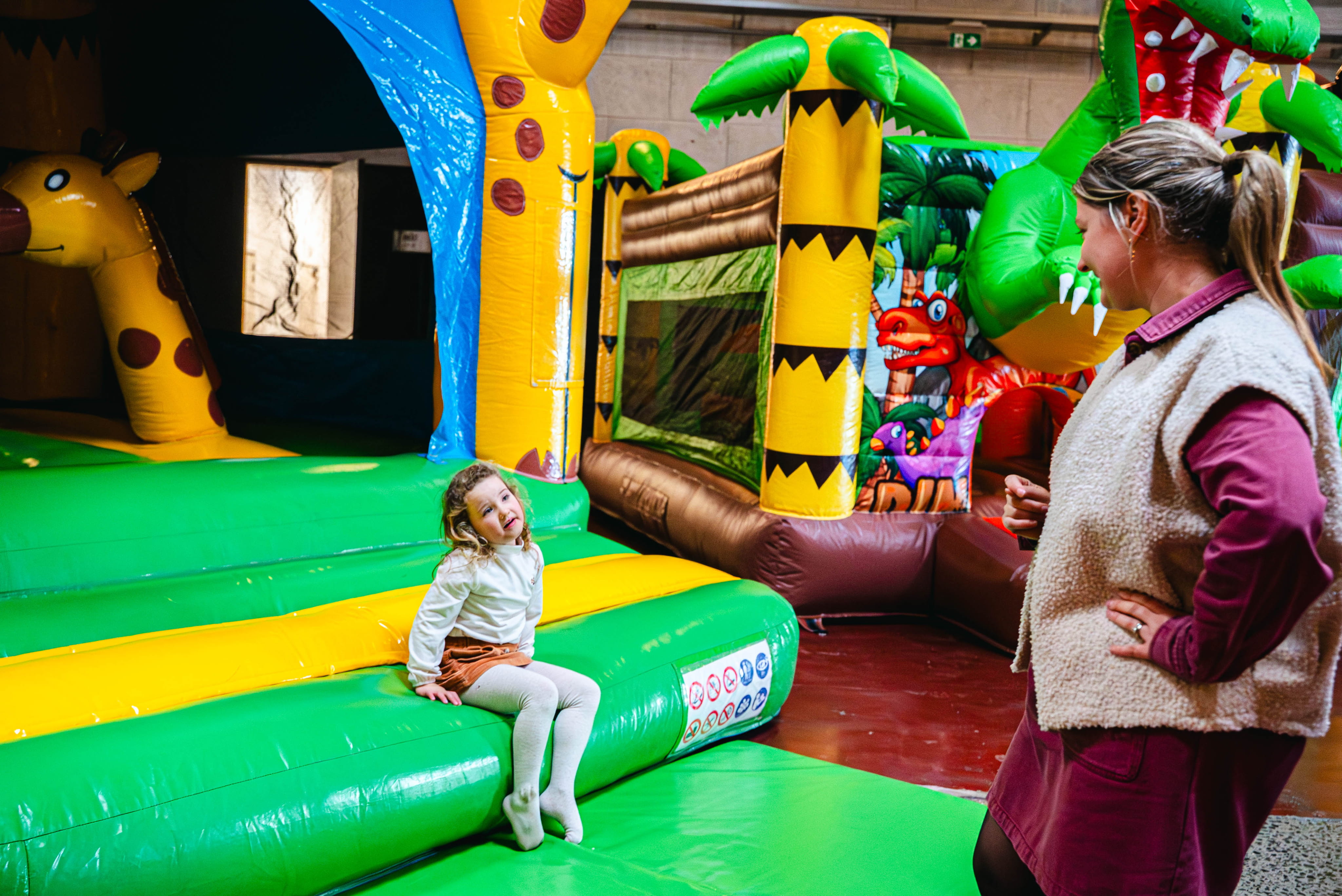 Een jong meisje zit op de rand van een jungle-thema springkasteel met felle kleuren en dierenprints, terwijl een lachende volwassene toekijkt. Het springkasteel is opgezet in een binnenruimte.