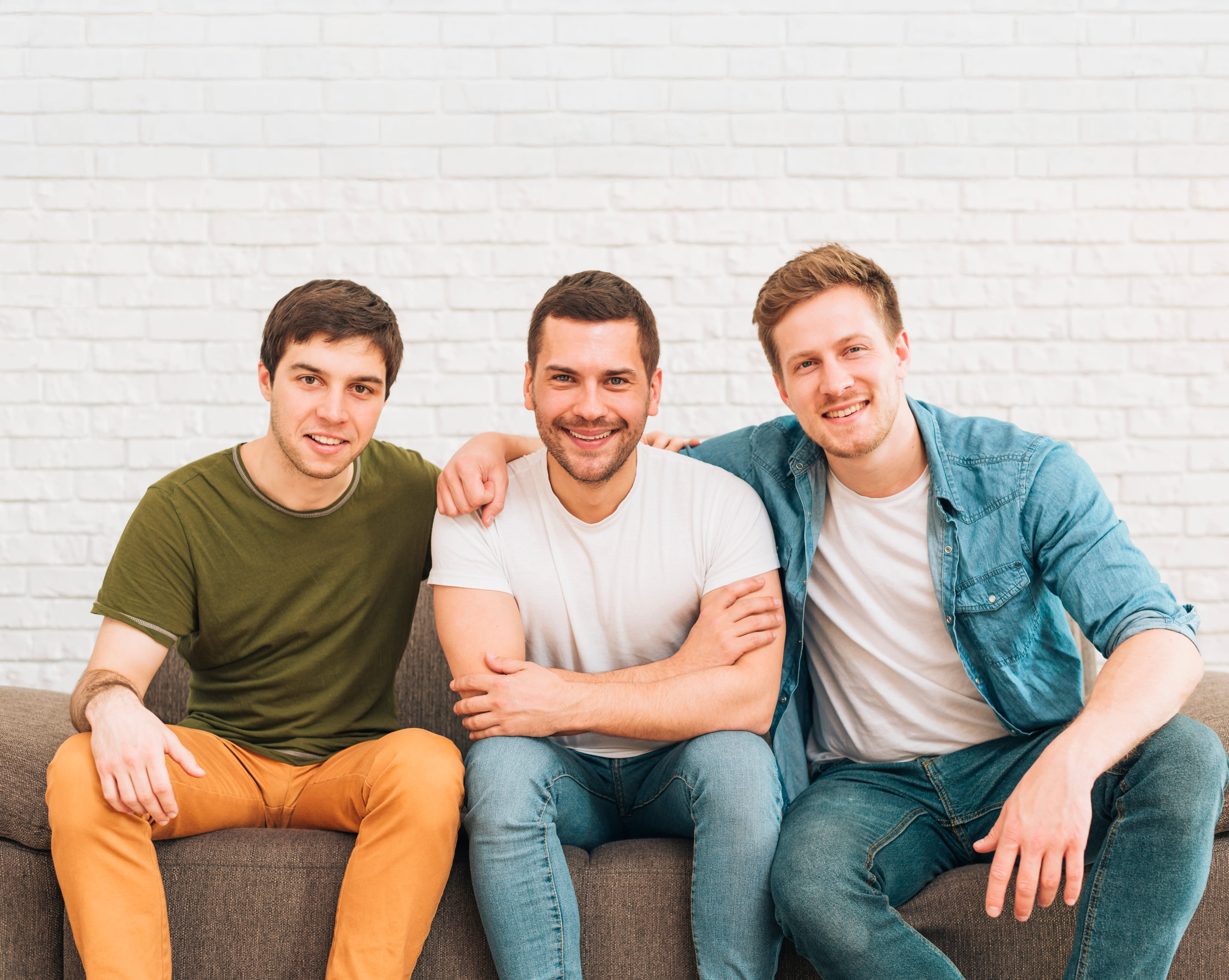 Drie jonge volwassenen zitten naast elkaar op een zetel en lachen naar de camera, met hun armen ontspannen om elkaar heen, tegen een witte bakstenen achtergrond.