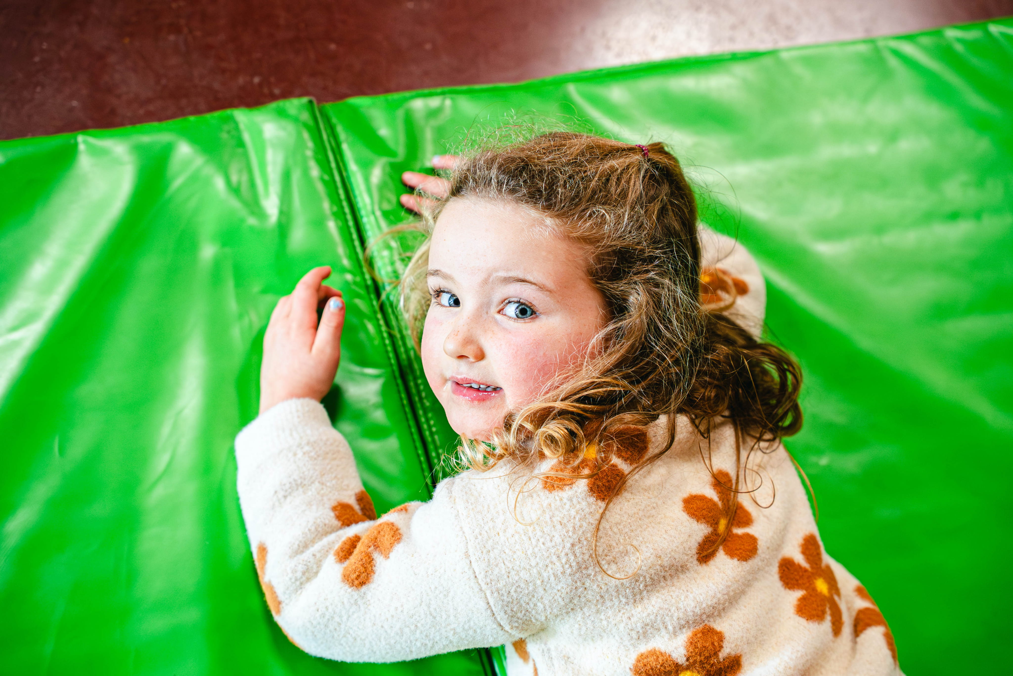 Een jong meisje met krullend haar ligt op haar buik op een felgroen springkasteel en kijkt lachend om zich heen.