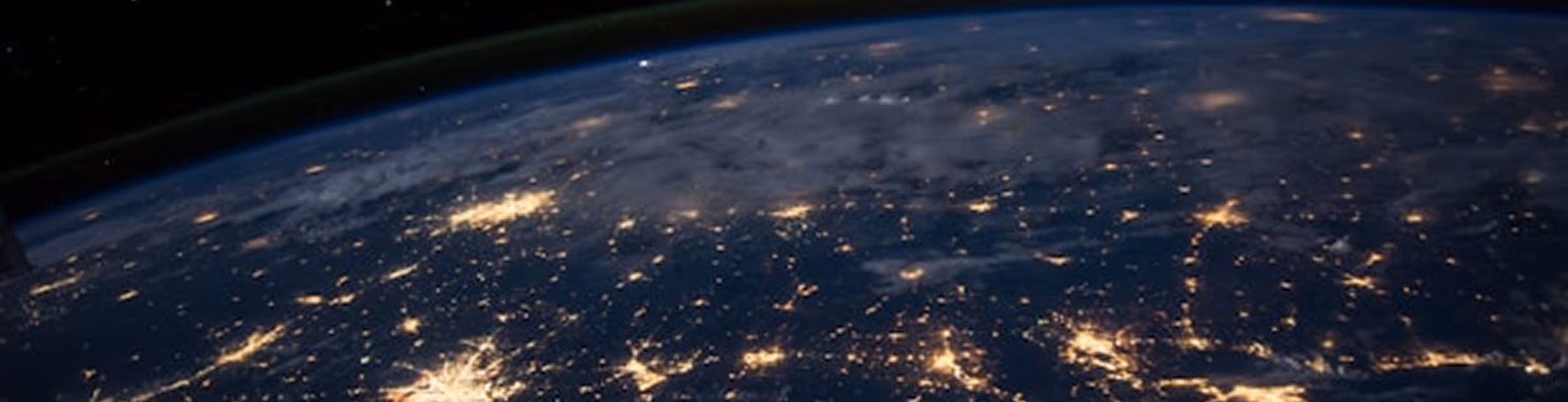 View of Earth's surface at night from space showing illuminated city lights and dark clouds against the dark outer space background.