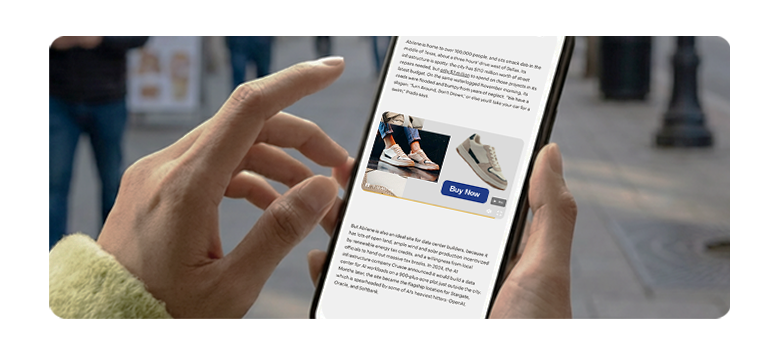 Close-up of a person holding a smartphone in both hands, viewing an online shopping app displaying two sneakers with prices and a blue 'Buy Now' button on a city street background.