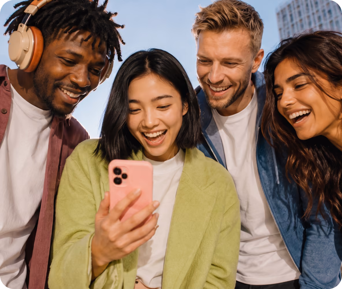 Four diverse young adults smiling and laughing as they look at a pink smartphone held by the woman in a green jacket outdoors.