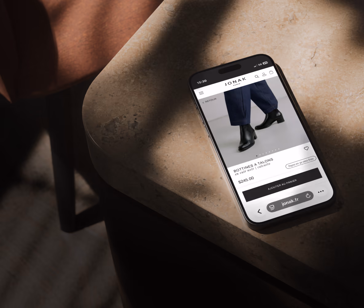 Smartphone on a stone table displaying a product page for black heeled ankle boots priced at $245 on the Jonak website.