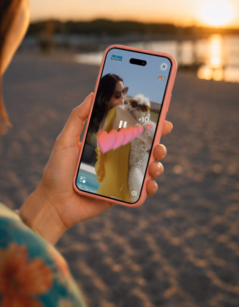 Hand holding phone displaying video of woman in sunglasses holding a dog wearing sunglasses on beach.