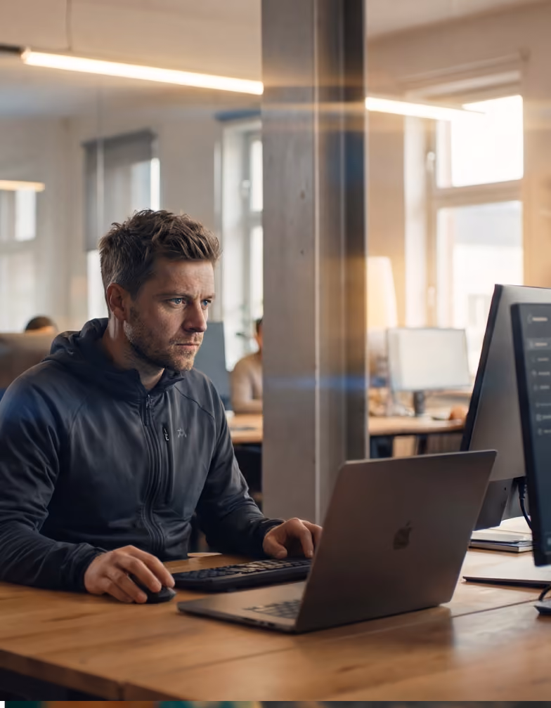 Man in a dark hoodie working intently on a laptop with a desktop monitor in a bright office.