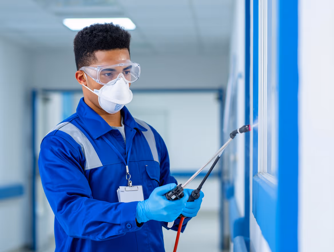 Un technicien en combinaison bleue, portant un masque, des lunettes de protection et des gants, désinfecte une surface avec un pulvérisateur dans un couloir.