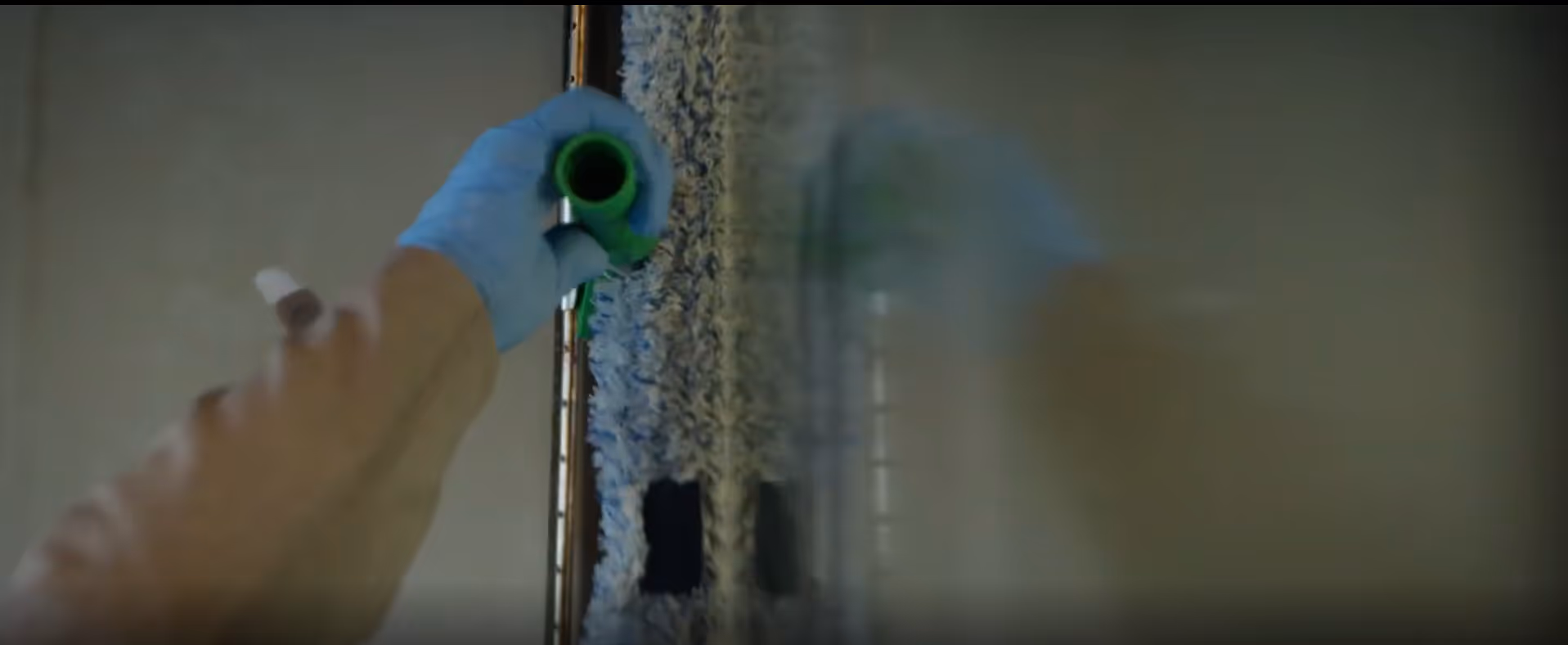 Une main gantée bleue tient un outil vert pour nettoyer une surface vitrée avec une raclette.