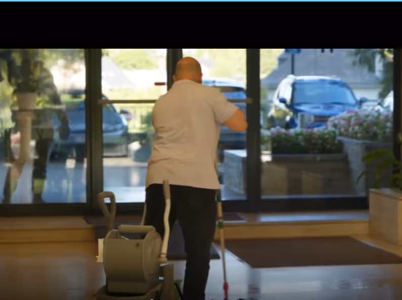 Un homme de dos utilise un balai mécanique dans un hall d'entrée avec des portes vitrées et des voitures visibles à l'extérieur.