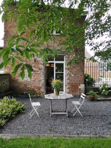 Table ronde en bois avec un vase de fleurs, entourée de deux chaises blanches sur une terrasse en gravier devant une maison en briques.
