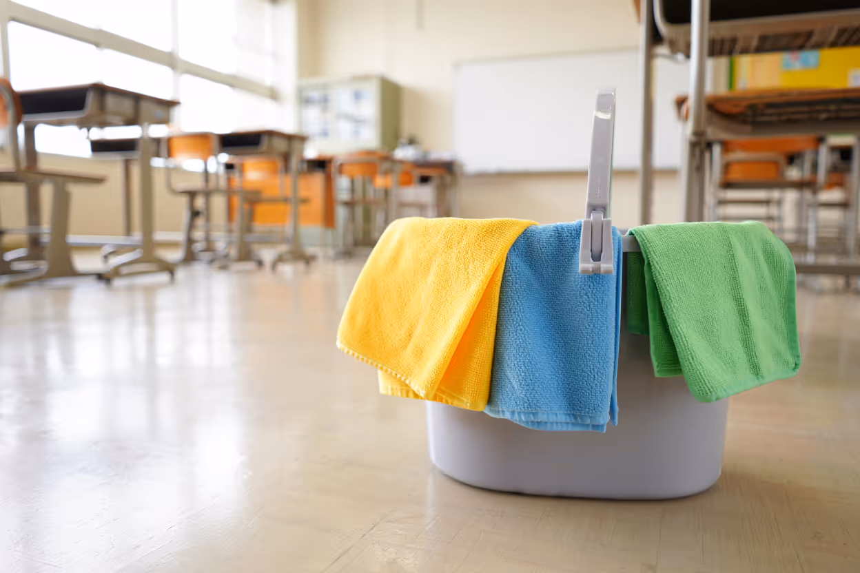 Seau de nettoyage avec chiffons jaune, bleu et vert posé sur un sol de salle de classe vide.