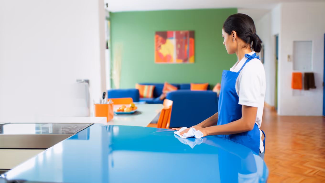 Femme en tablier bleu nettoyant un plan de travail bleu dans une cuisine moderne avec un salon coloré à l'arrière-plan.