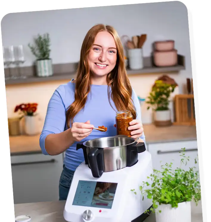 Smiling young woman in a kitchen adding sauce from a jar into a food processor on the counter.