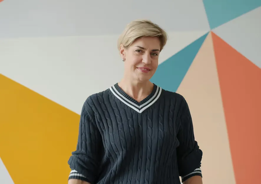 Smiling woman with short blonde hair wearing a dark blue sweater with white stripes, standing in front of a colorful geometric wall.