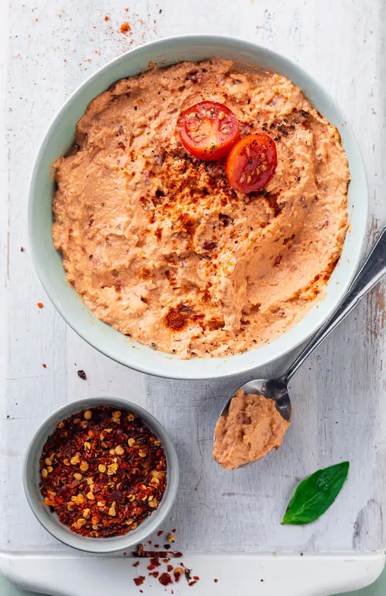 Bowl of creamy hummus garnished with two cherry tomato halves and sprinkled with chili flakes, next to a spoonful of hummus and a small bowl of crushed chili flakes on a white wooden surface.
