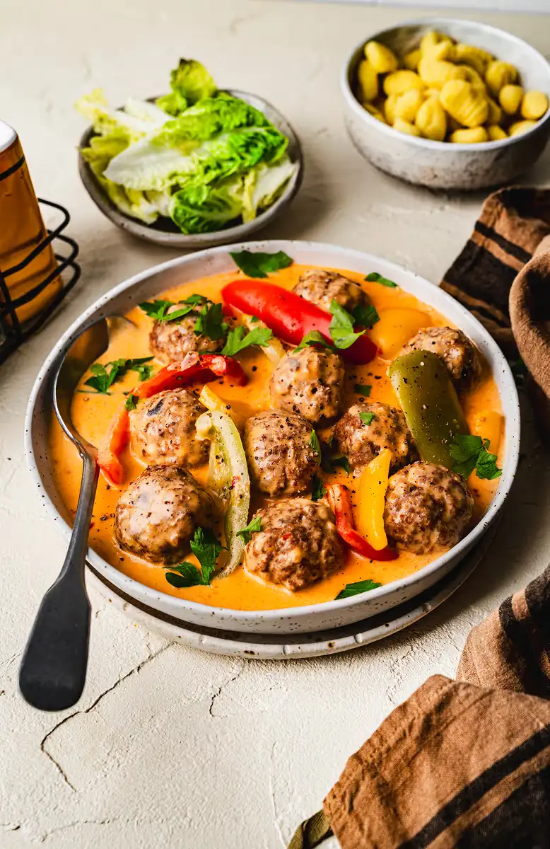 Plate of meatballs in creamy orange sauce with bell peppers and parsley, accompanied by bowls of lettuce and gnocchi.
