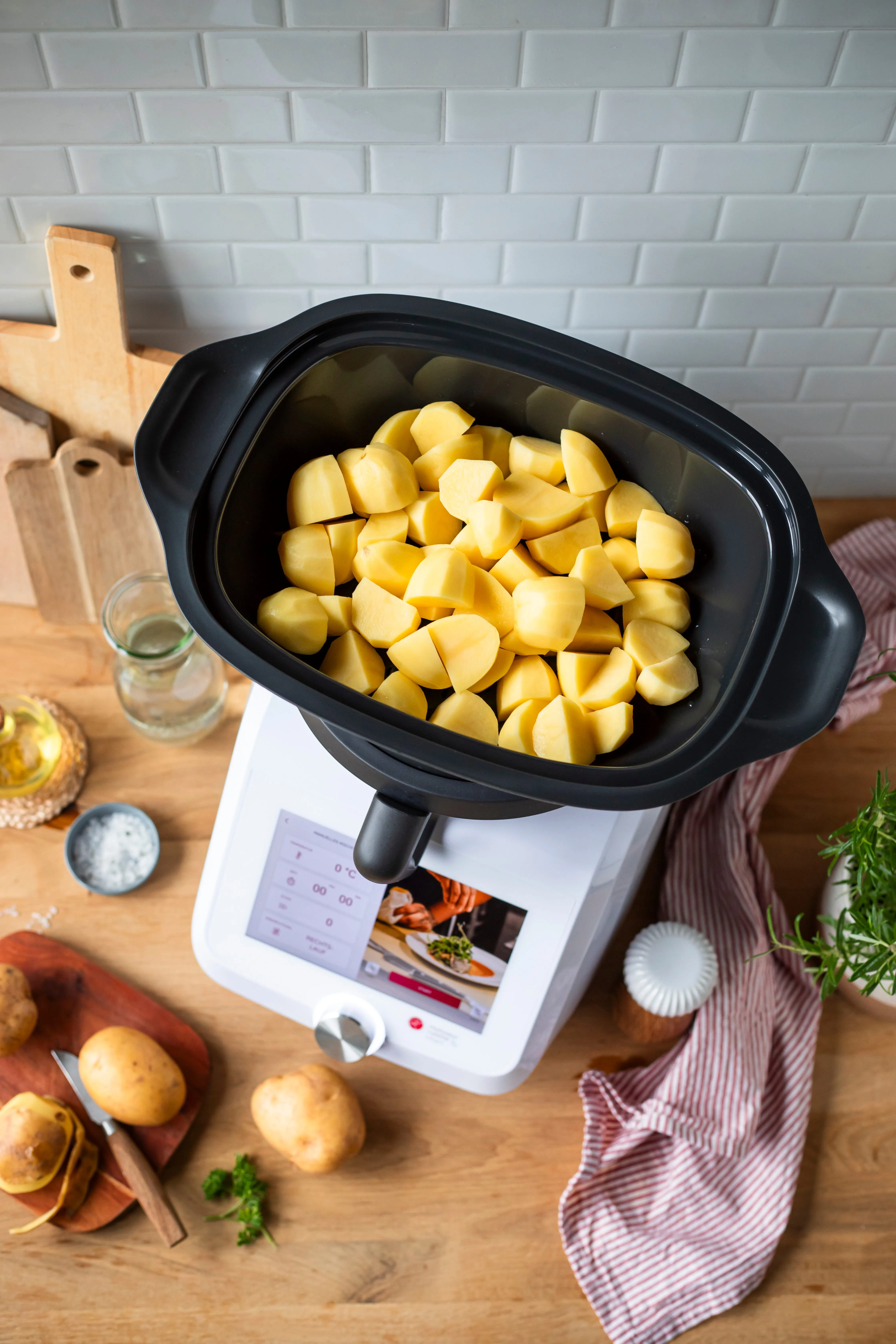 Smart kitchen appliance with peeled and chopped potatoes inside a black container on a wooden countertop with potatoes, knife, cutting boards, herbs, and striped cloth around.