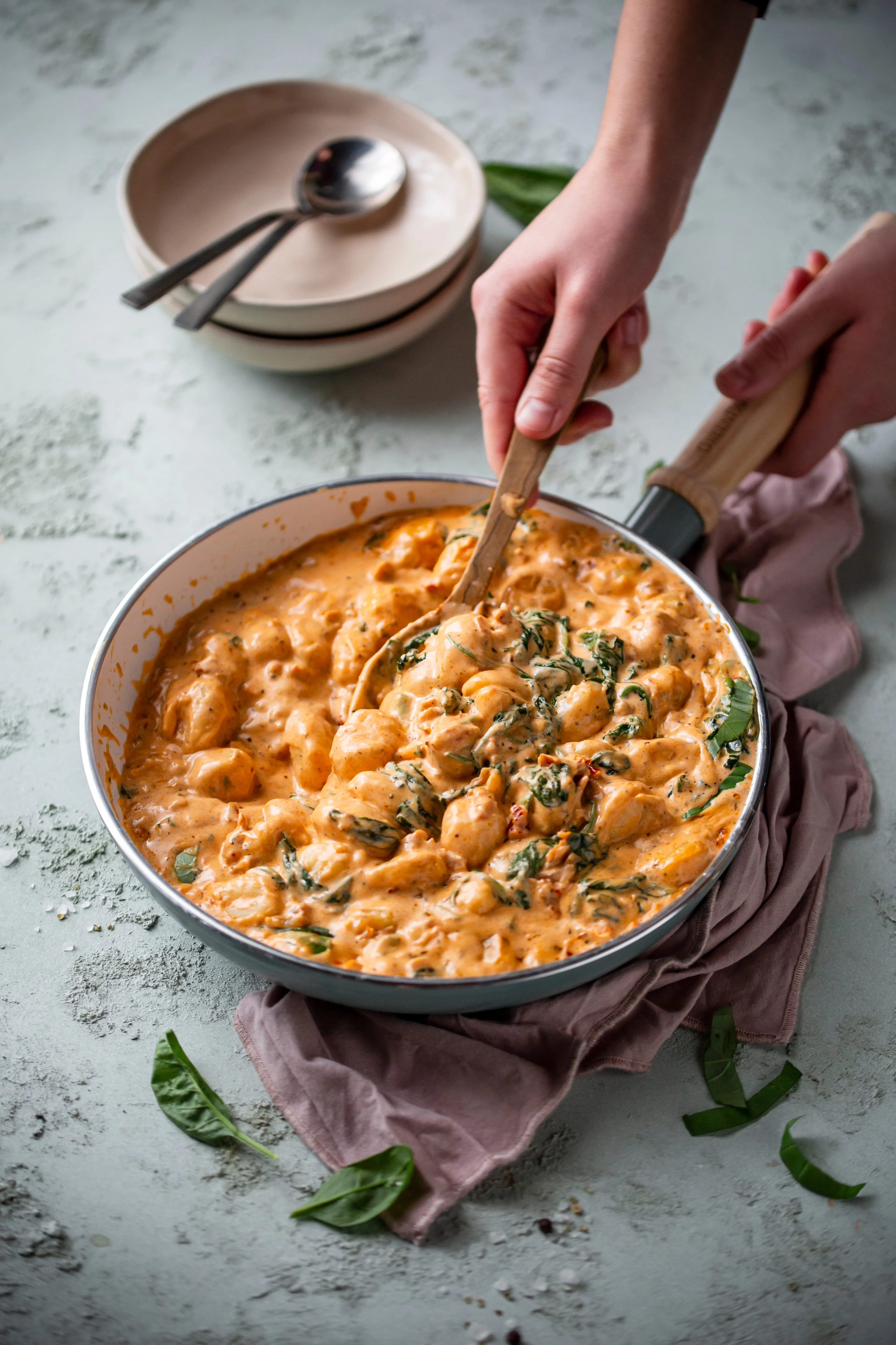 Hands stirring creamy gnocchi with spinach in a skillet on a purple cloth.
