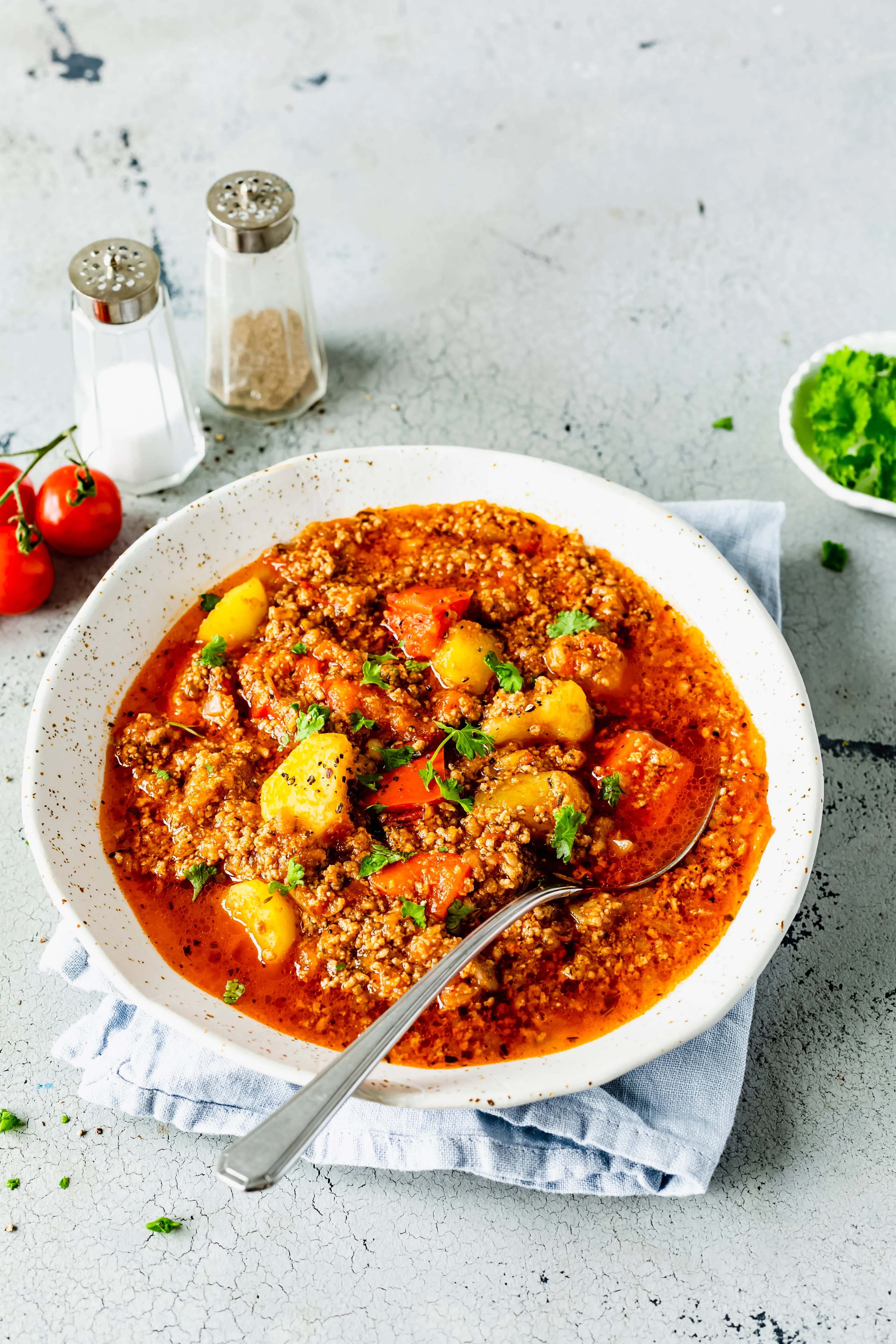 Bowl of hearty ground meat stew with potatoes, carrots, and parsley on a light cloth napkin beside salt and pepper shakers and cherry tomatoes.