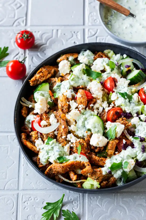 Bowl of seasoned grilled meat strips with cucumber, cherry tomatoes, onions, feta cheese, fresh parsley, and creamy herb sauce.