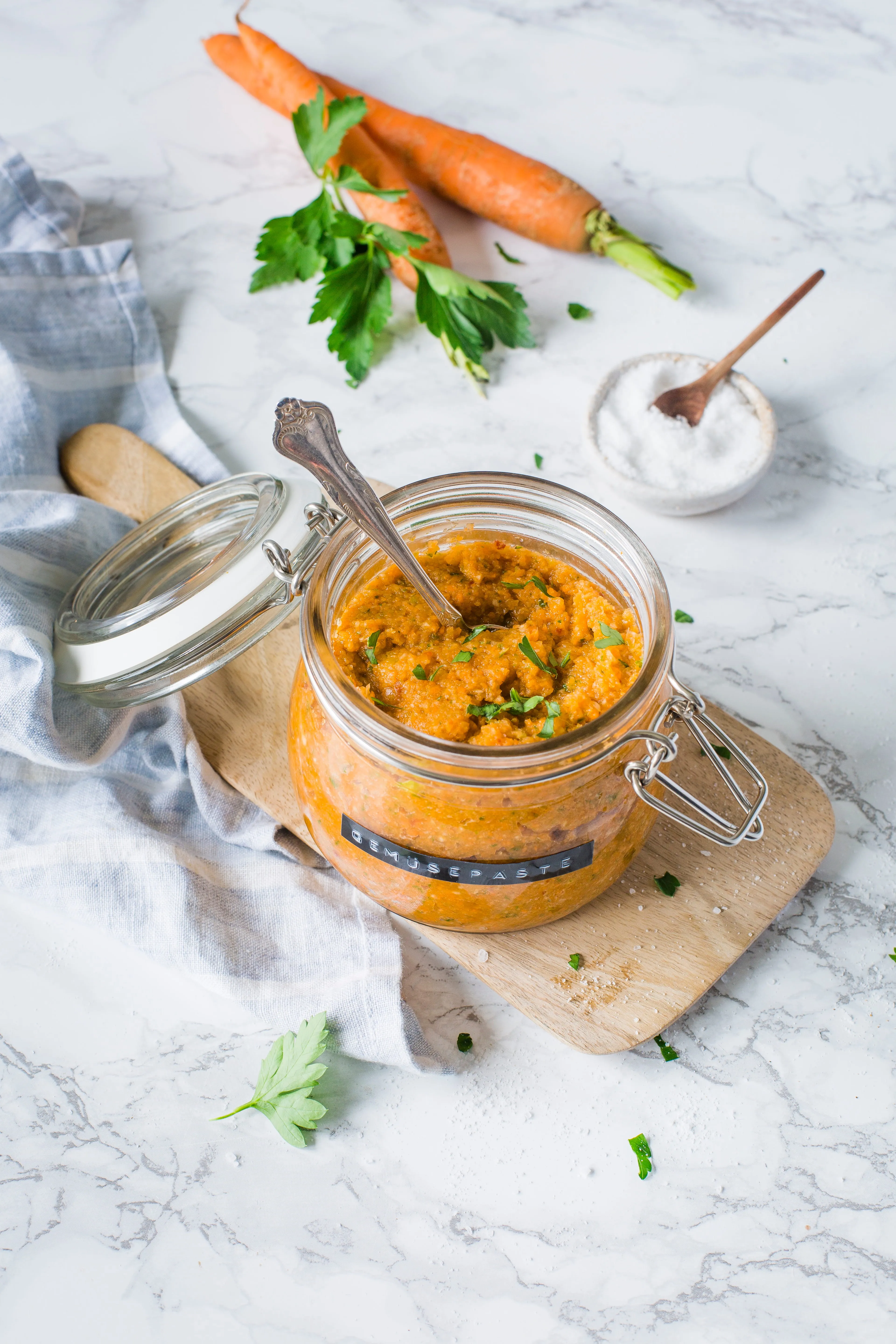 Glass jar of orange vegetable paste labeled 'Gemüsepaste' with a spoon inside, placed on a wooden board with two carrots and parsley leaves nearby on a marble surface.