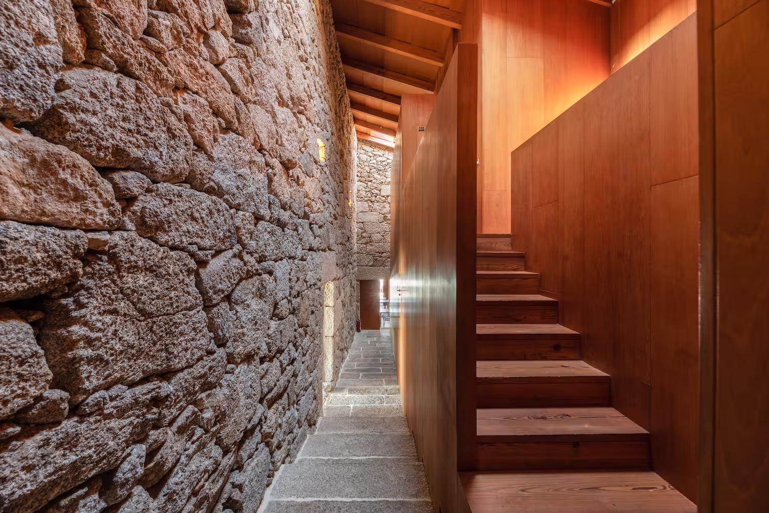 Narrow interior corridor with rough stone wall on the left and wooden stairs and paneling on the right under a wooden ceiling.