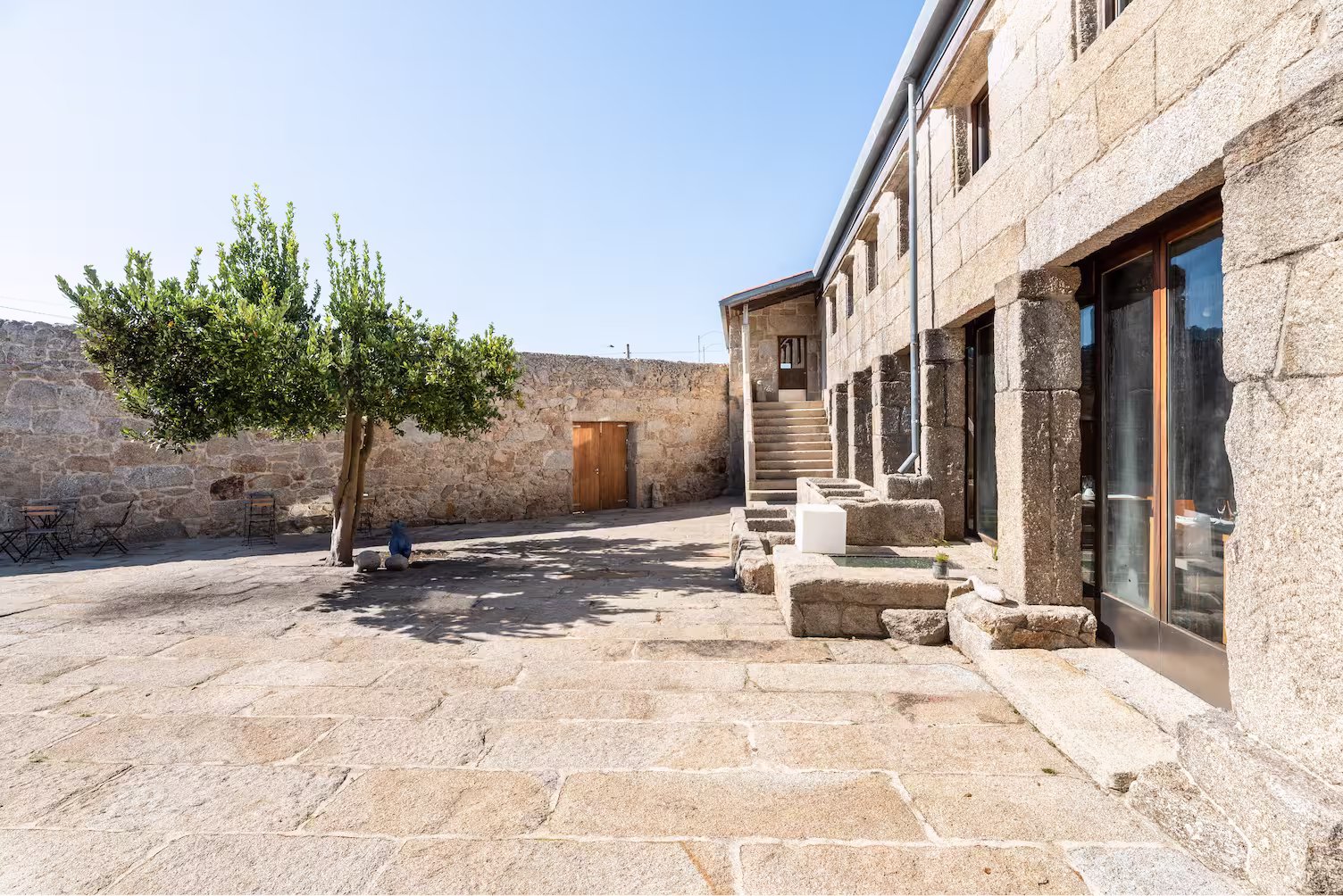 Sunny courtyard with stone flooring, a leafy tree, outdoor chairs, and a stone building with large windows and stairs.