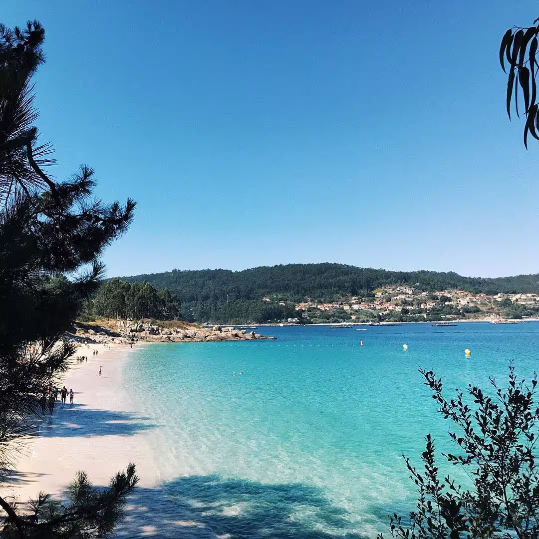 Clear turquoise water along a sandy beach with people walking, framed by trees and hills under a bright blue sky.