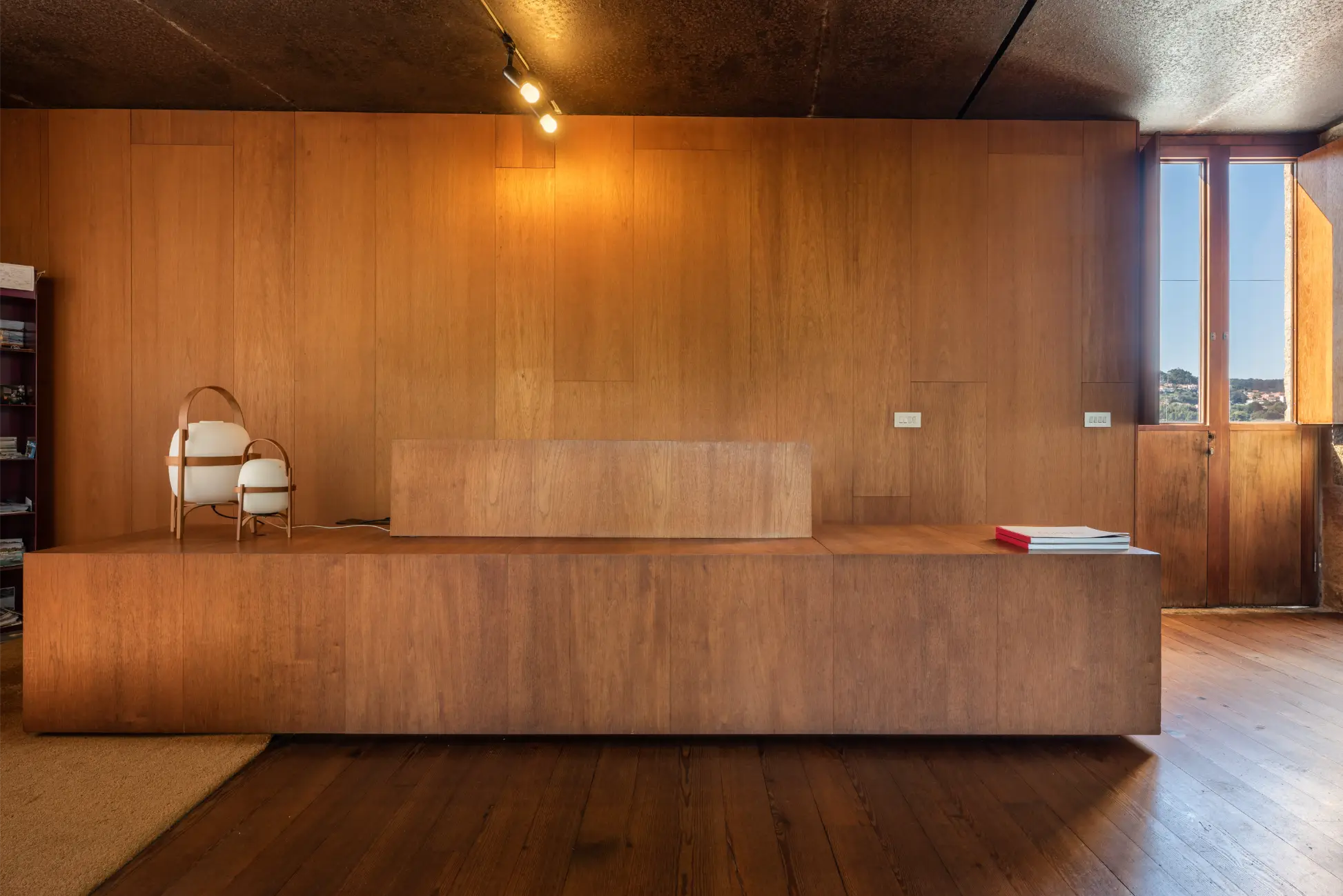 Minimalist wooden interior with built-in bench, two white lanterns on the left, and a stack of books near a window on the right.