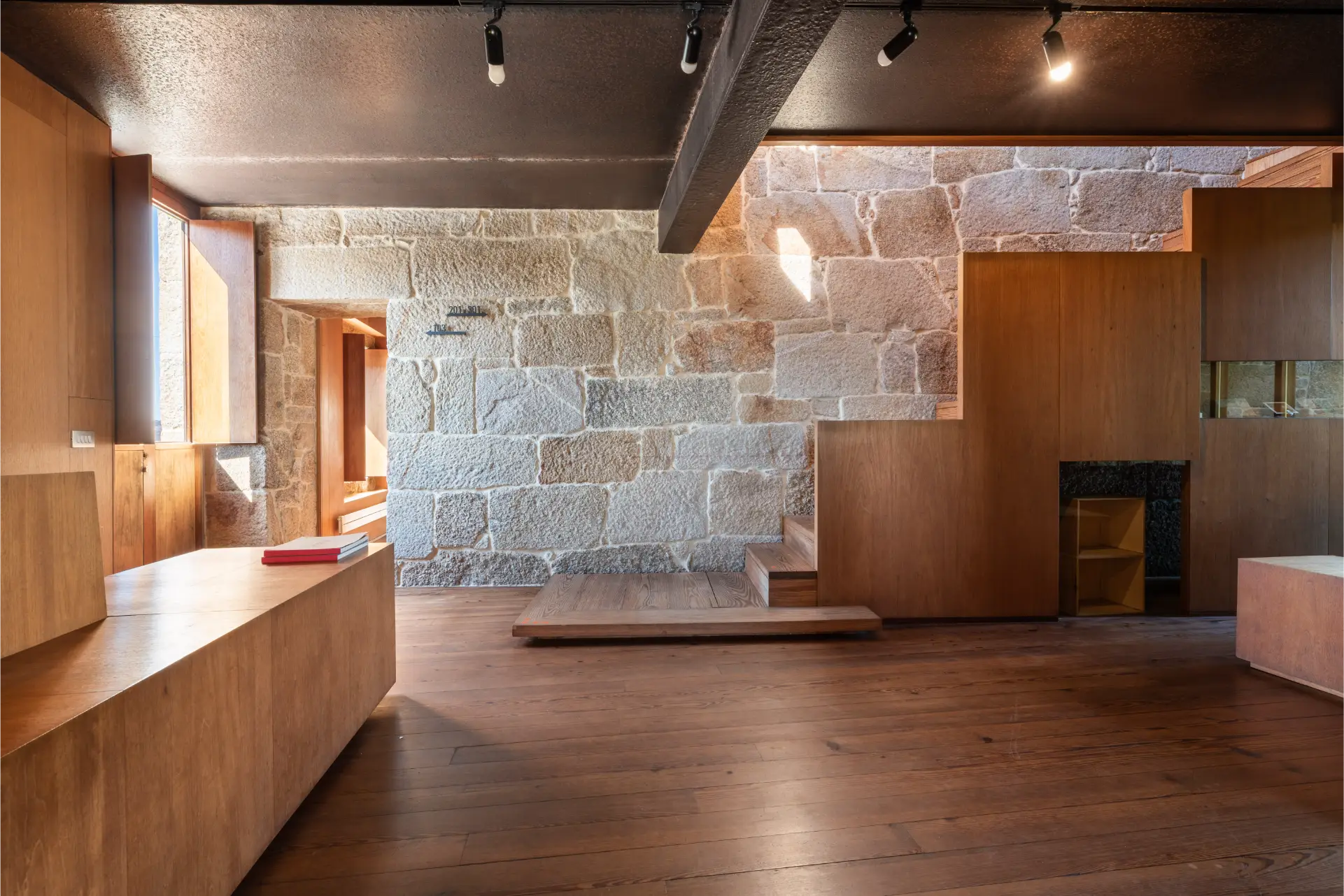 Modern interior with wooden flooring, furniture, and stairs against textured stone walls and large windows letting in natural light.