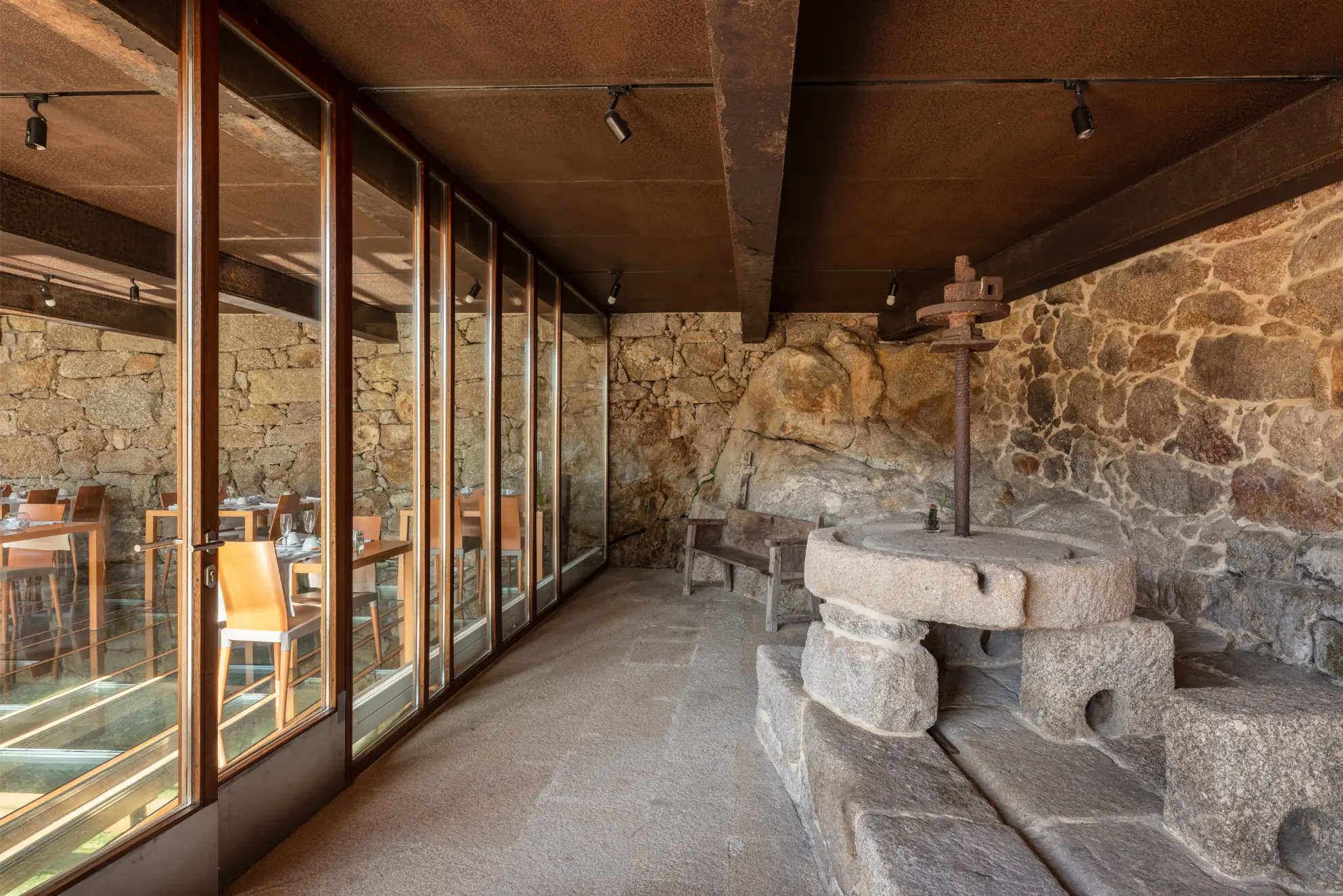 Interior of a rustic stone room with an old millstone, wooden bench, and glass doors reflecting a dining area with wooden chairs and tables set for a meal.