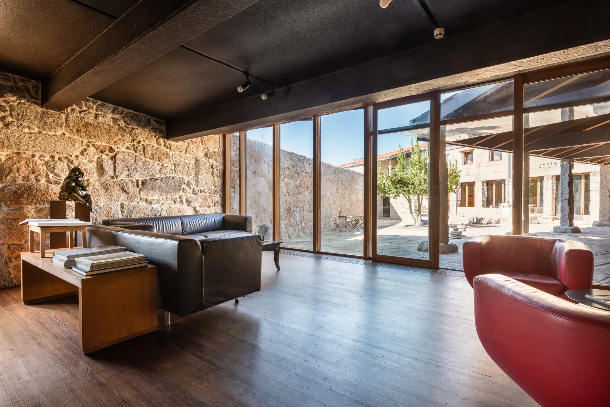 Modern living area with stone walls, black leather sofa, red armchair, wooden coffee table, and large glass doors overlooking a stone courtyard with a tree.