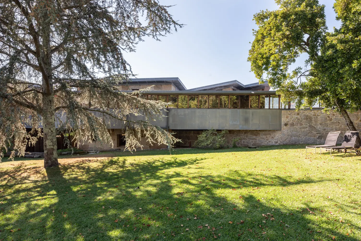 Modern house with stone walls and large windows, surrounded by green lawn and shaded by tall trees.