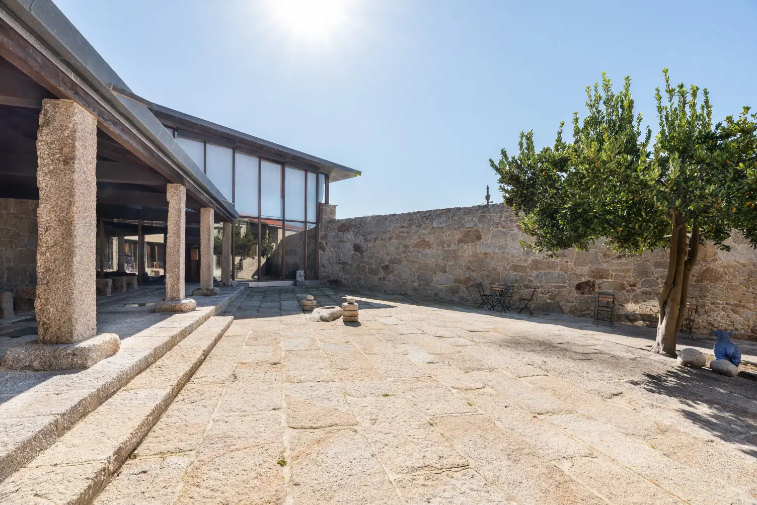 Sunlit stone courtyard with pillars on the left, a citrus tree on the right, and a stone wall in the background under a clear blue sky.