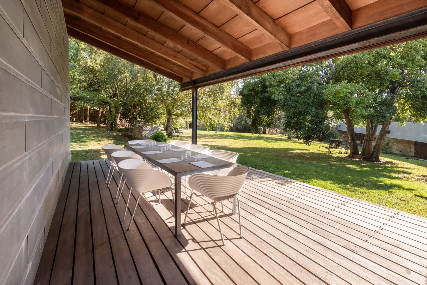 Outdoor wooden deck with a long table and eight white chairs under a wooden roof, overlooking a green grassy yard with trees.