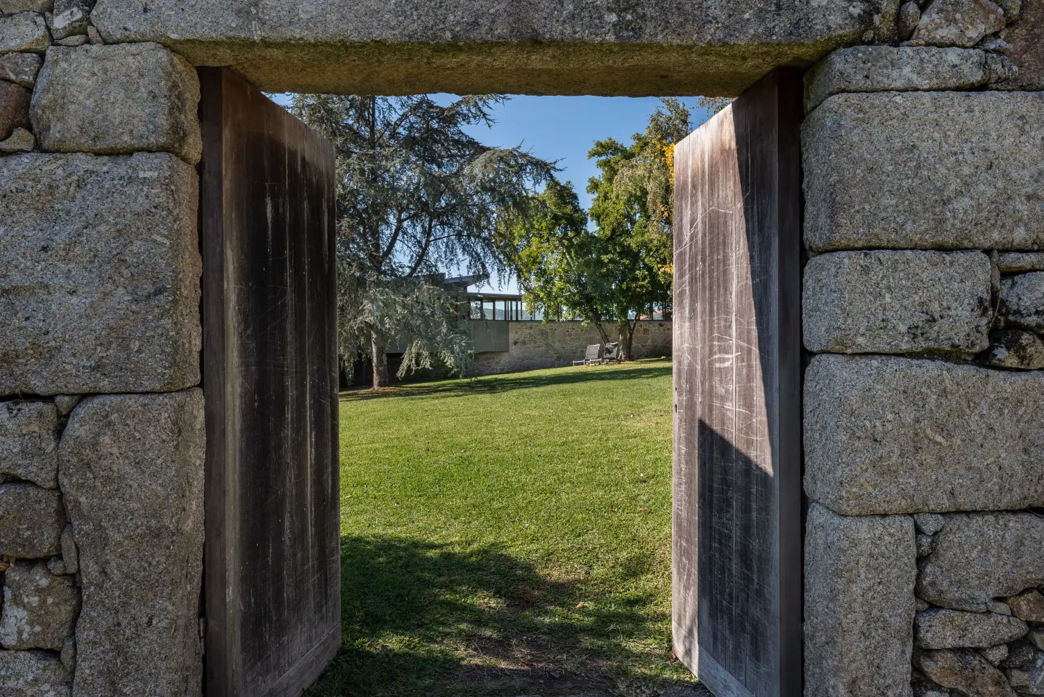 Open wooden doors set in a stone wall reveal a green lawn with trees and two lounge chairs under a clear blue sky.