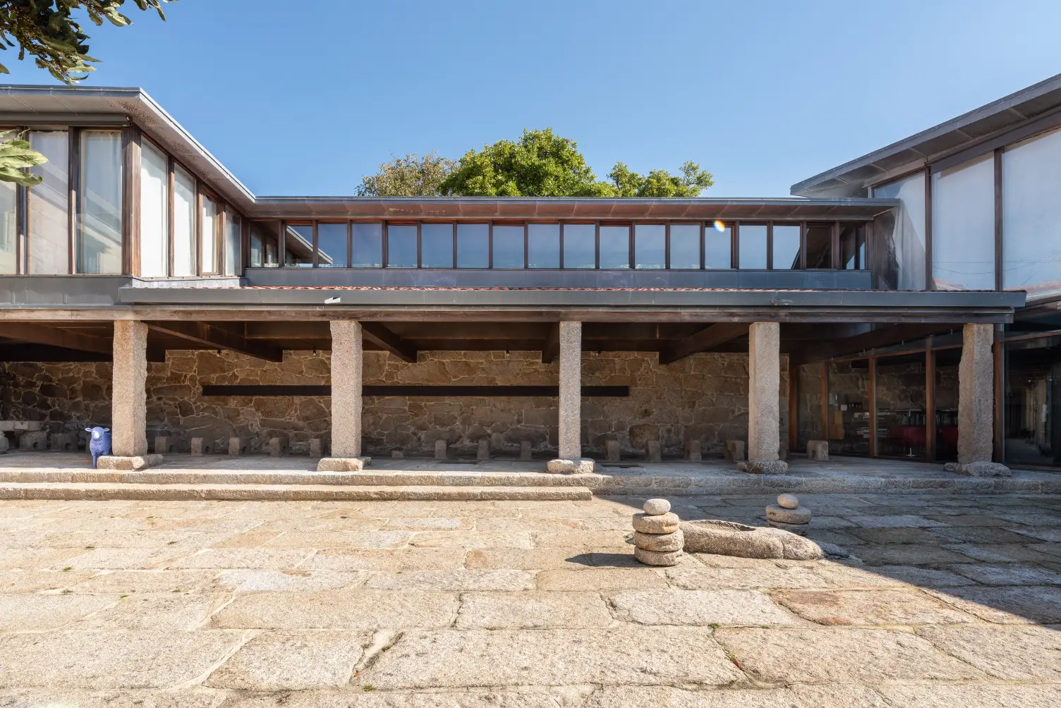 Stone courtyard with columns supporting a modern glass and stone building under a clear blue sky.
